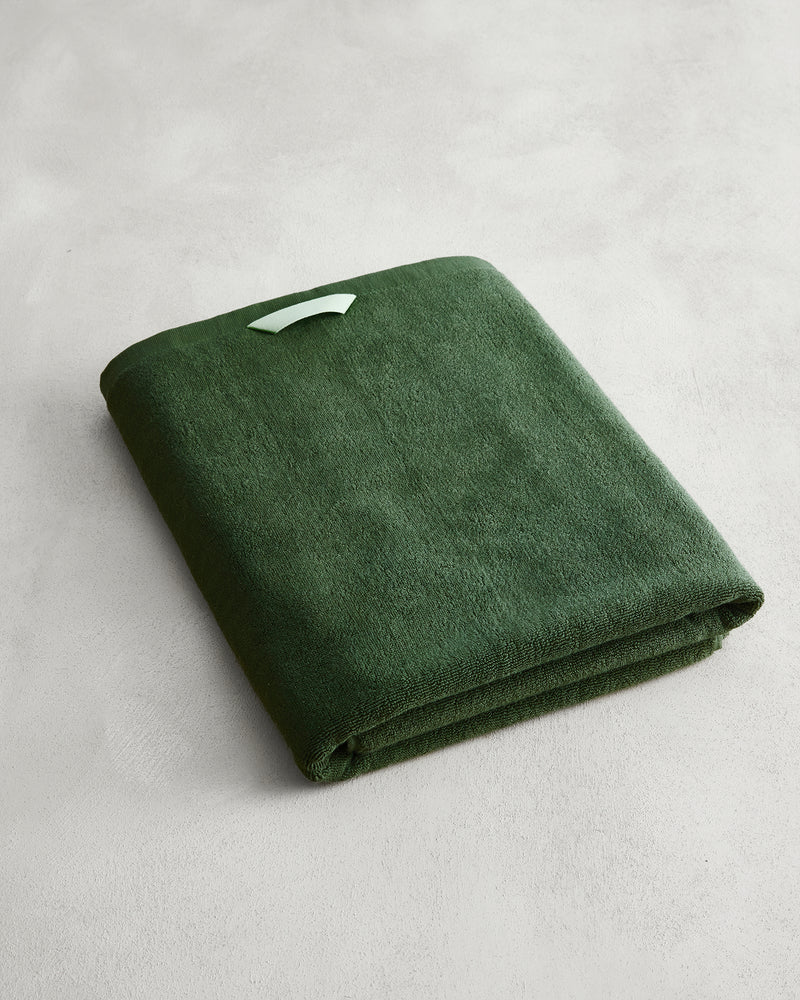 The Moss 100% Cotton Terry Bath Sheet by Bed Threads, in deep green with a white tag, rests on a light gray surface—ideal for anyone seeking plush, quick-drying towels.