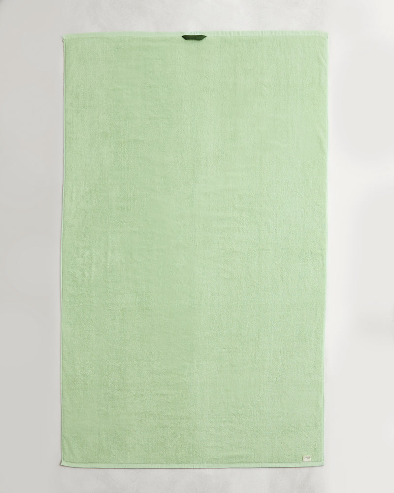 A set of two Apple 100% Cotton Terry Bath Sheets by Bed Threads, in light green, each rectangular with a dark green loop near the top edge and a white tag at the bottom right, laid flat on a white surface.