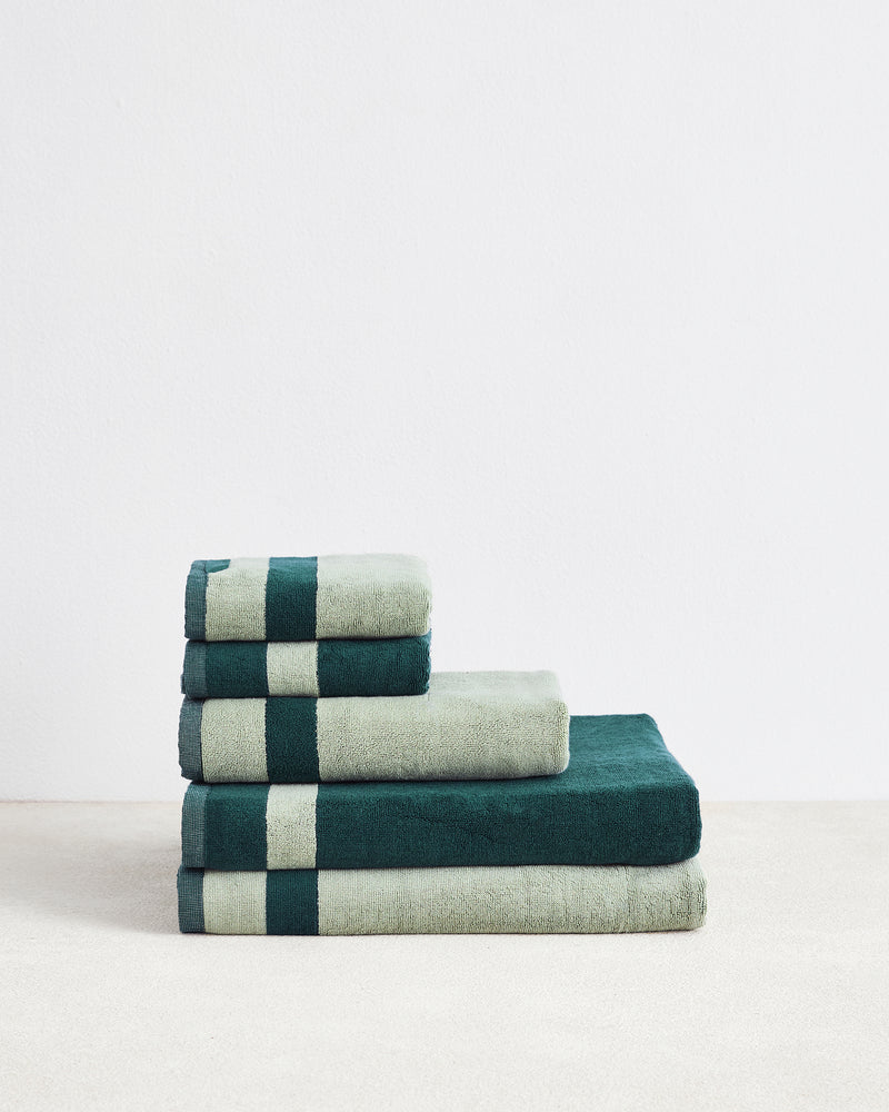 A stack of Bed Threads’ Forest & Sage Two-Toned 100% Cotton Terry Bath Sheet Bundle, arranged from largest to smallest, sits against a white wall on a light surface.