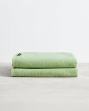 A set of two neatly folded Apple 100% Cotton Terry Bath Sheets by Bed Threads is shown on a light gray surface against a plain white background, each featuring a small tag. - view