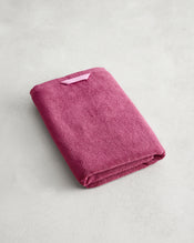 A folded Berry 100% Cotton Terry Bath Towel by Bed Threads, deep pink with a small loop on one corner, rests on a light gray textured surface. - view