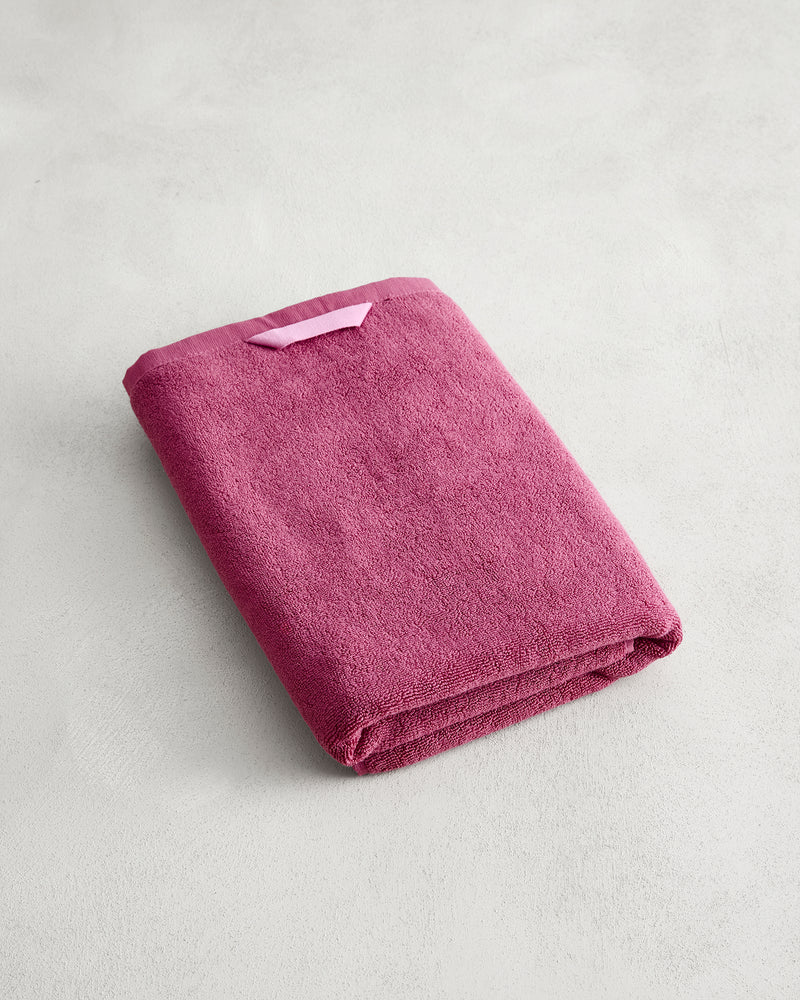 A folded Berry 100% Cotton Terry Bath Towel by Bed Threads, deep pink with a small loop on one corner, rests on a light gray textured surface.