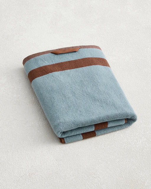 The Bed Threads Cacao & Lagoon Two-Toned 100% Cotton Terry Bath Towel, featuring blue and brown stripes plus a small brown tag, is folded on a light textured surface.