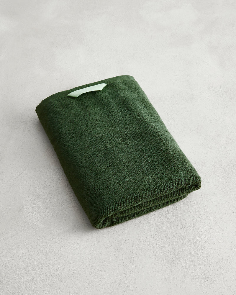A folded Moss 100% Cotton Terry Bath Towel by Bed Threads in dark green rests on a light grey textured surface, with a small light green tag attached to its corner.