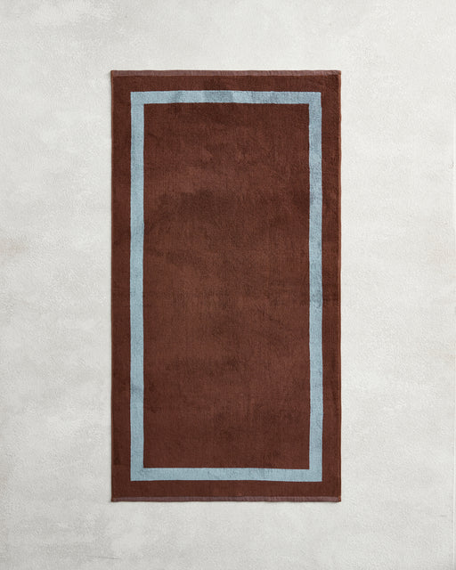 The Cacao & Lagoon Two-Toned 100% Cotton Terry Bath Towel by Bed Threads adds a cozy touch when paired with a brown rug featuring a light blue border, creating an inviting vibe for your bathroom.