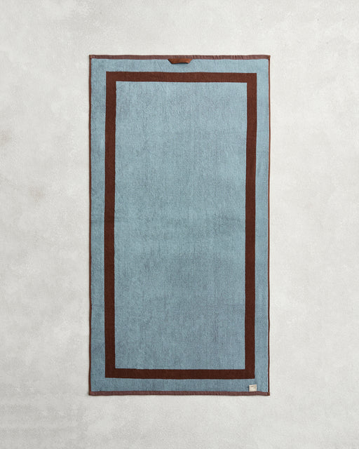 The Bed Threads Cacao & Lagoon Two-Toned 100% Cotton Terry Bath Towel Bundle is shown laid flat, featuring a blue rectangular center, dark brown border with an inner brown frame, and a small tag on the bottom right corner.