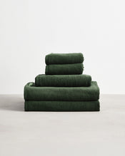 A neatly stacked Bed Threads Moss 100% Cotton Terry Bath Towel Bundle in dark green, featuring various sizes, is arranged on a light gray surface against a plain white background. - view