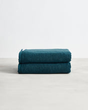 A set of two Aegean 100% Cotton Terry Bath Towels by Bed Threads rests folded on a light gray surface against a white background, highlighting their plush, highly absorbent texture. - view
