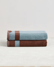 Two Cacao & Lagoon Two-Toned 100% Cotton Terry Bath Towels by Bed Threads are stacked on a light surface, showcasing their plush texture and modern design—one features blue and brown stripes, the other is brown with a blue stripe. - view