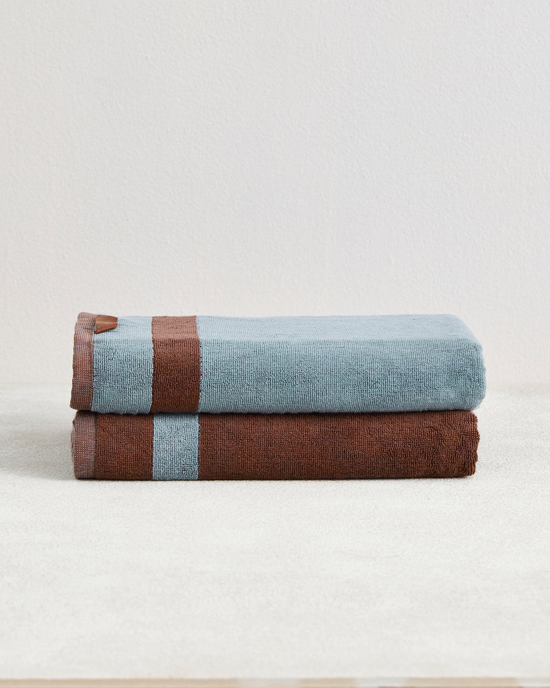 Two Cacao & Lagoon Two-Toned 100% Cotton Terry Bath Towels by Bed Threads are stacked on a light surface, showcasing their plush texture and modern design—one features blue and brown stripes, the other is brown with a blue stripe.