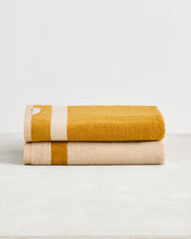 Two Crème & Turmeric Two-Toned 100% Cotton Terry Bath Towels by Bed Threads are stacked on a light surface—one mustard yellow with a beige stripe, the other beige with a mustard yellow stripe—adding style to your bathroom. - view