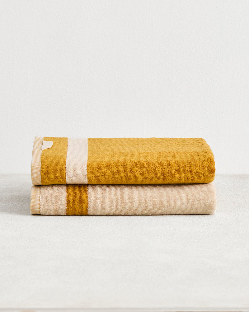 Two Crème & Turmeric Two-Toned 100% Cotton Terry Bath Towels by Bed Threads are stacked on a light surface—one mustard yellow with a beige stripe, the other beige with a mustard yellow stripe—adding style to your bathroom.