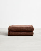 A set of two Bed Threads Espresso 100% Cotton Terry Bath Towels, neatly folded with a small beige tag, rests on a light gray surface against a white background. - view