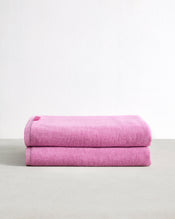 A set of two Bubblegum 100% Cotton Terry Bath Sheets from Bed Threads, plush and highly absorbent in pink, is displayed folded on a light gray surface with a plain white background. - view