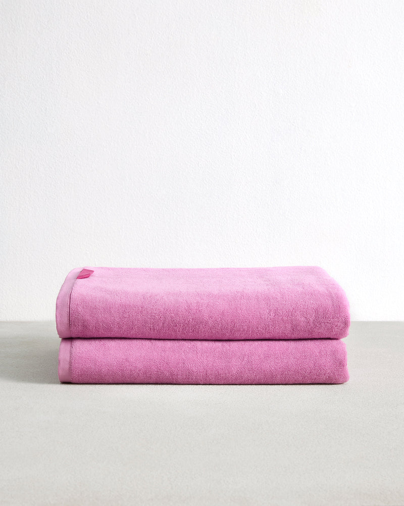 A set of two Bubblegum 100% Cotton Terry Bath Sheets from Bed Threads, plush and highly absorbent in pink, is displayed folded on a light gray surface with a plain white background.