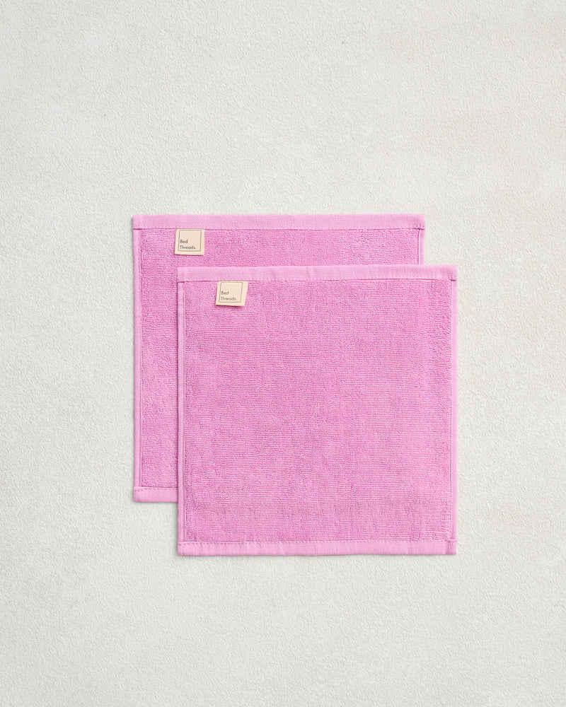 Two Bubblegum 100% Cotton Terry Face Washers by Bed Threads are displayed on a light gray surface, slightly overlapping. Their soft, absorbent terry fabric is gentle on skin. Set includes two rectangular pink face washers with light pink tags.