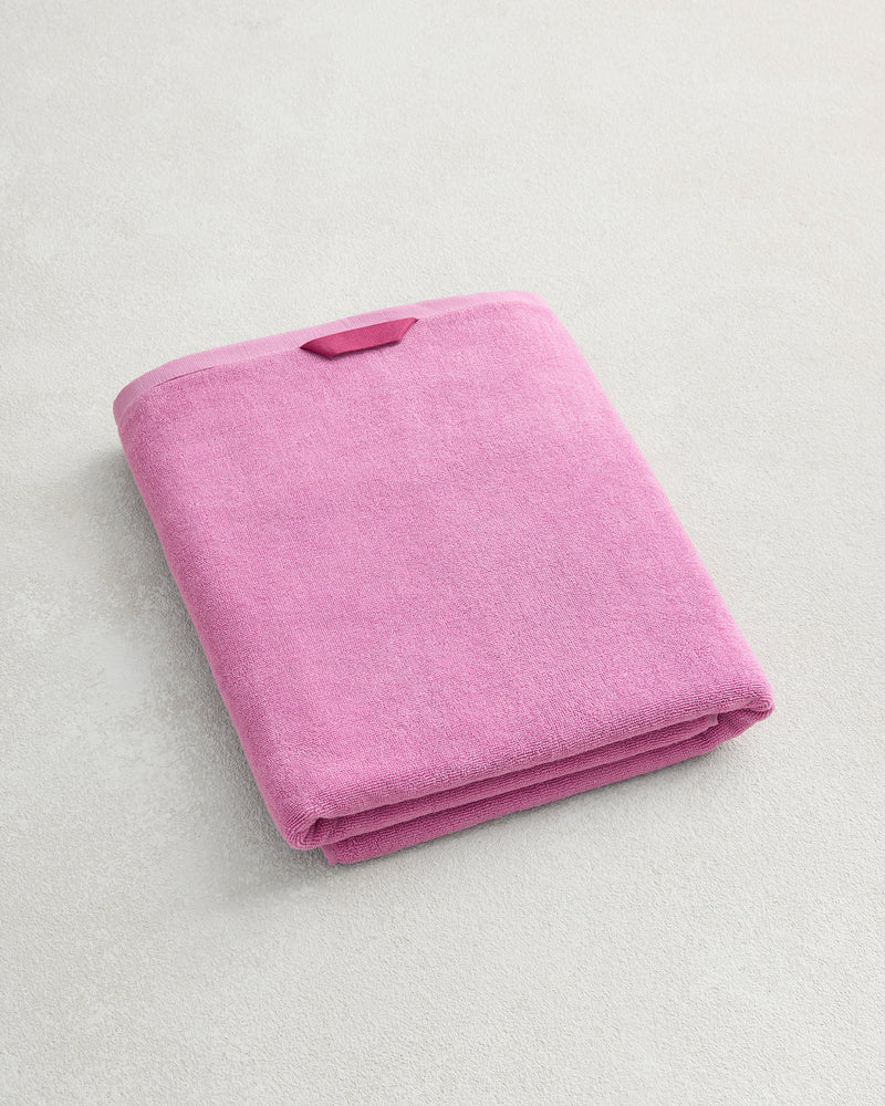 A Bubblegum 100% Cotton Terry Bath Sheet by Bed Threads, plush and quick-drying, with a small rectangular tag, sits neatly folded on a light-colored textured surface.