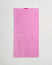The Bed Threads Bubblegum 100% Cotton Terry Bath Towel, made from quick-drying 650GSM terry, lies flat on a light textured surface. It features finished edges and a small hanging loop near the top. - view