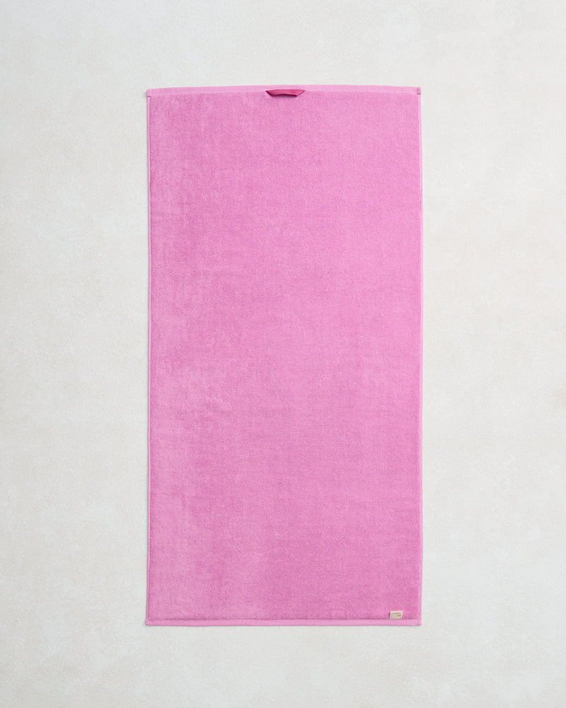The Bed Threads Bubblegum 100% Cotton Terry Bath Towel, made from quick-drying 650GSM terry, lies flat on a light textured surface. It features finished edges and a small hanging loop near the top.