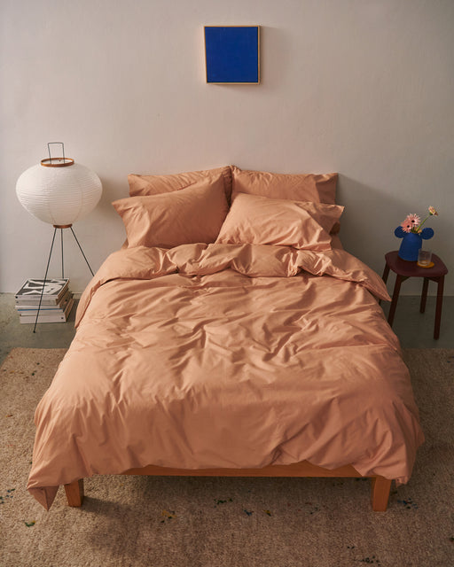 A neatly made bed with light brown bedding and Bed Threads' Butterscotch Organic Cotton King Pillowcases stands against a pale wall. To the left is a round white floor lamp and stacked books; to the right, a small table with pink flowers in a vase. A blue square artwork hangs above.