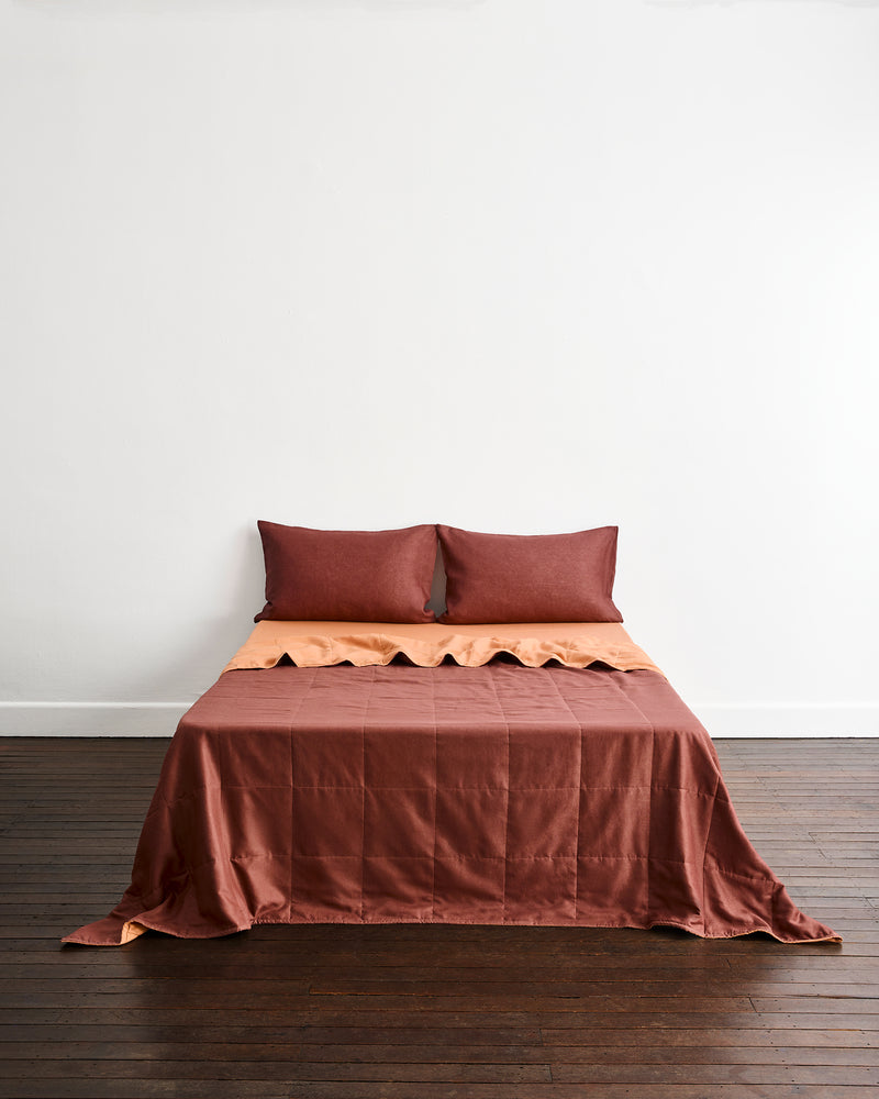 A bed dressed in a rust-brown quilt cover in Cacao & Hazelnut with matching pillowcases in Cacao, set against a plain white wall and dark wood floor.