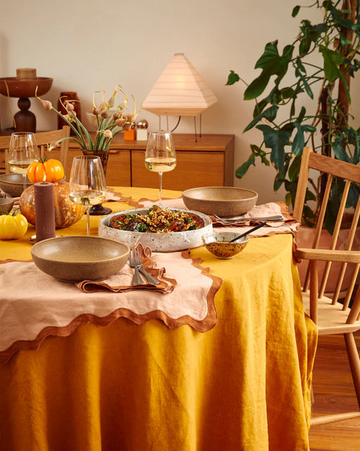 A dining table set for a meal features Bed Threads’ Terracotta & Rust 100% French Flax Linen Scalloped Napkins, plates, bowls, wine glasses, pumpkins, candles, and a salad bowl with wooden chairs in a cozy, warmly lit room.