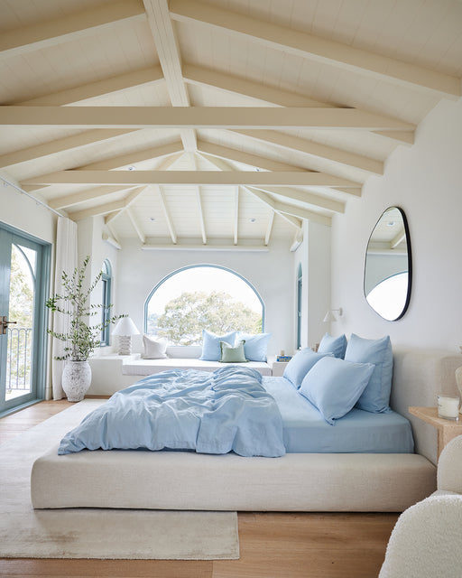 A bright bedroom with vaulted white ceilings, a large arched window, beige bed with the Coast 100% French Flax Linen Duvet Cover from Bed Threads, a round mirror, plants, and natural light create a serene, airy atmosphere.