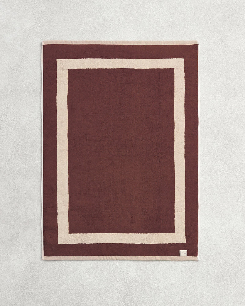 Flat cotton terry bath mat on a grey background in a cacao colour with a creme border.