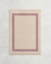 Flat cotton terry bath mat on a grey background in a creme colour with a lavender border. - view