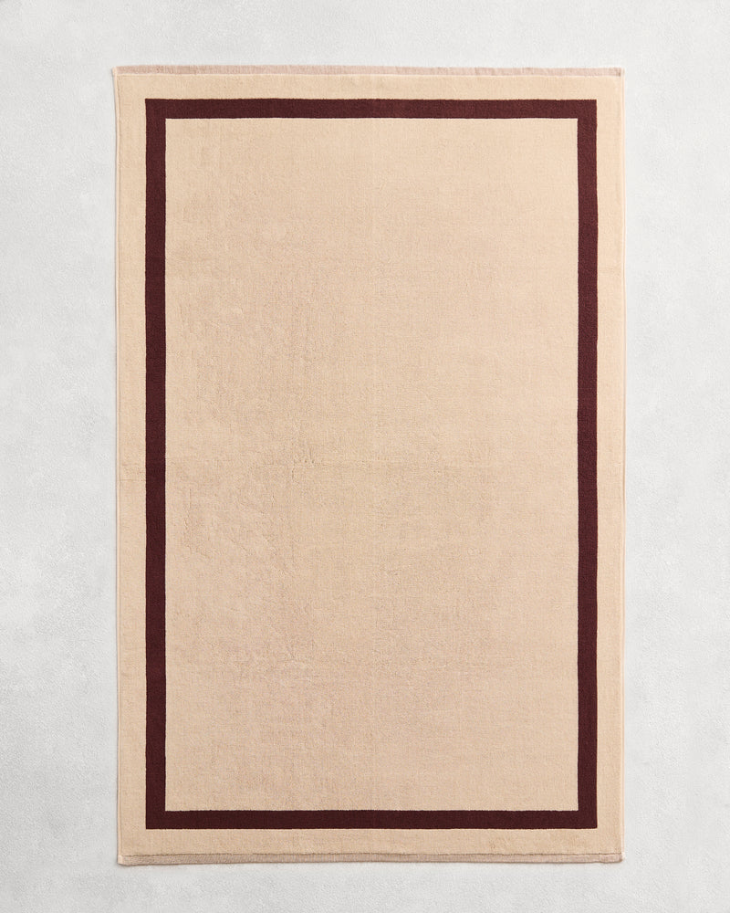 Flat cotton terry bath sheet on a grey background in a creme colour with a cacao border.