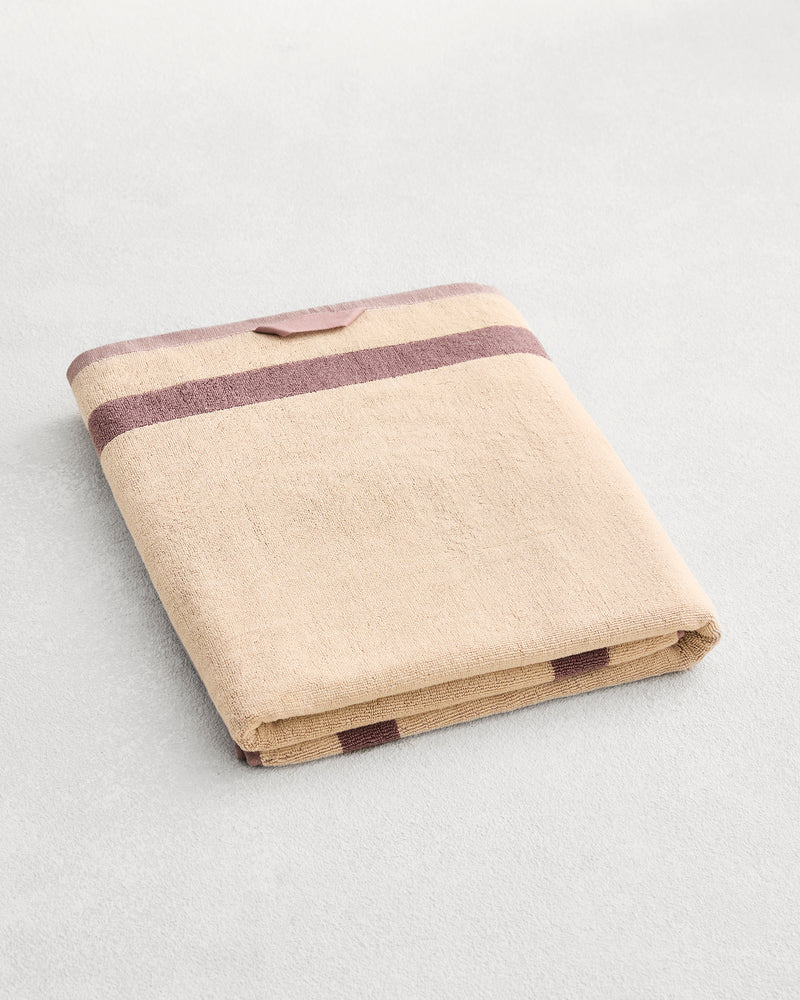 A folded cotton terry bath sheet on a grey background, in a creme colourway with a lavender border.