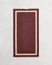 Flat cotton terry bath towel on a grey background in a cacao colour with a creme border. - view