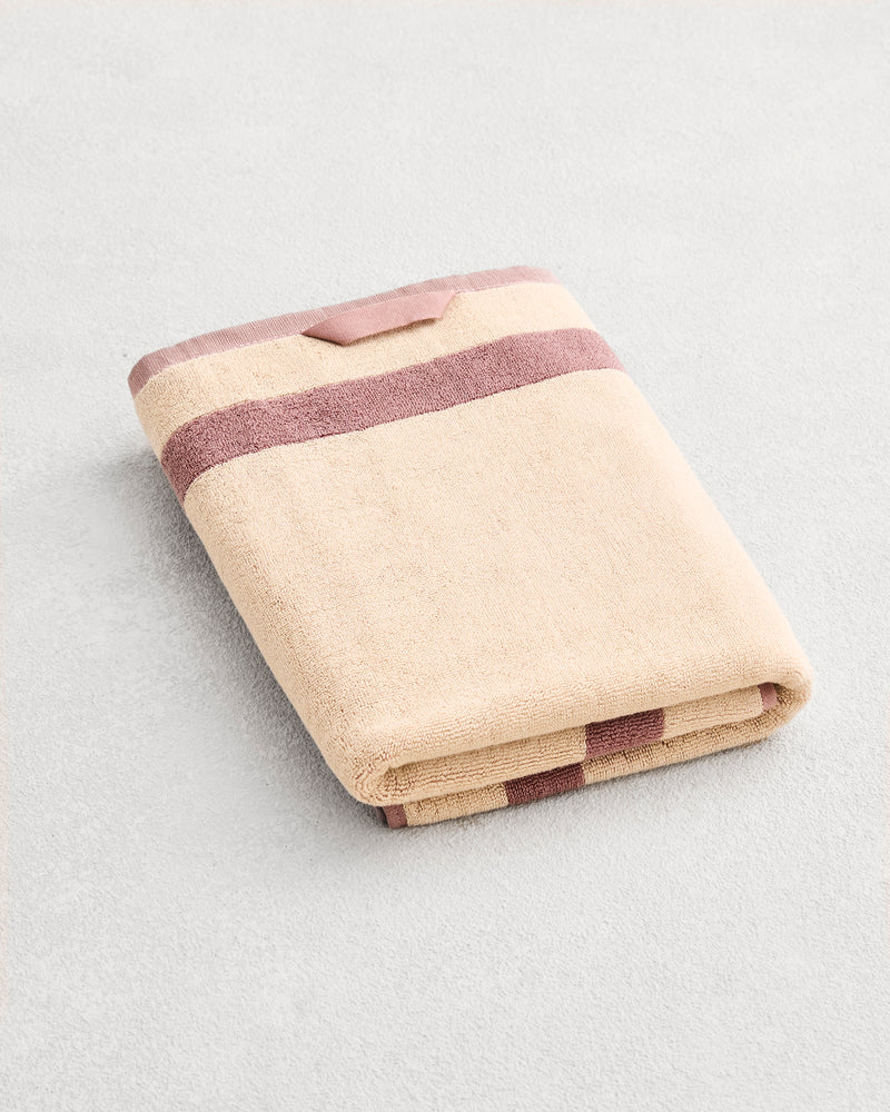 A folded cotton terry bath towel on a grey background, in a creme colourway with a lavender border.