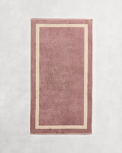 Flat cotton terry bath towel on a grey background in a lavender colour with a creme border. - view