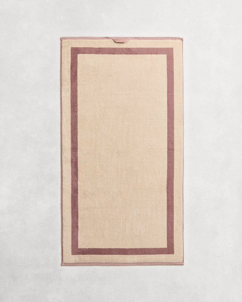 Flat cotton terry bath towel on a grey background in a creme colour with a lavender border.