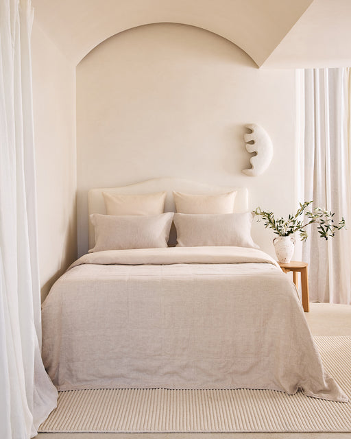 A cozy, minimalist bedroom features a neatly made bed with Bed Threads Crème 100% French Flax Linen European Pillowcases (Set of Two), two pillows, and neutral linens. Crescent wall decor and a vase of greenery sit on the wooden bedside table.