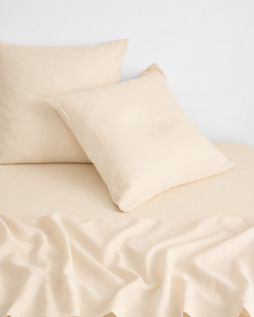 Two Bed Threads Crème 100% French Flax Linen European Pillowcases (Set of Two) rest on a matching bedsheet, with part of the sheet softly draped in the foreground against a plain white wall.