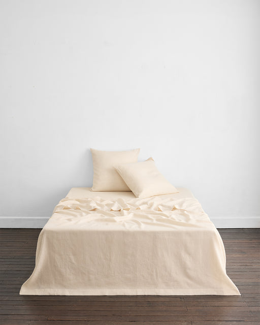 A neatly made bed features Bed Threads Crème 100% French Flax Linen European Pillowcases (Set of Two) with matching pillows, set against a white wall on a dark wood floor. The soft, slightly crumpled linen creates a relaxed vibe.