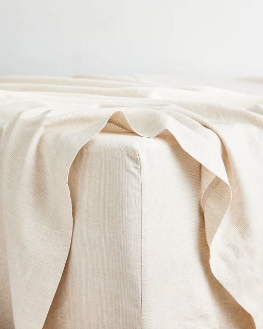 A close-up shows the Bed Threads Olive & Oatmeal & White Stripe 100% French Flax Linen Bedding Bundle, with the fitted sheet draped over the mattress corner. The minimal background highlights this cozy, premium bedding set.