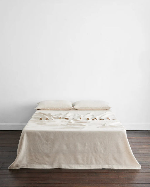 A minimalist bed featuring the Bed Threads Olive & Oatmeal & White Stripe 100% French Flax Linen Bedding Bundle sits against a white wall on a dark wooden floor, with two cream pillows and distinctive central triangular folds in the bedding.
