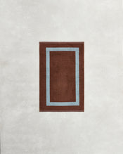 A rectangular brown rug with a light blue double border sits on a textured background, perfectly coordinating with Bed Threads' Cacao & Lagoon Two-Toned 100% Cotton Terry Hand Towels for a stylish bathroom ensemble. - view