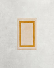 A rectangular beige rug with a mustard border rests on a light grey surface, reflecting the plush 650GSM softness and two-toned style of Bed Threads' Crème & Turmeric 100% Cotton Terry Hand Towel. - view