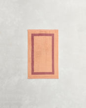 A rectangular peach bath mat with a darker peach border lies on a light gray surface, perfectly complementing Bed Threads’ Terracotta & Pink Clay Two-Toned 100% Cotton Terry Hand Towel. - view
