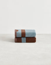 Two Bed Threads Cacao & Lagoon Two-Toned 100% Cotton Terry Hand Towels, featuring plush 650GSM fabric in light blue and brown, are neatly folded and stacked on a light surface against a plain white background. Set of two. - view