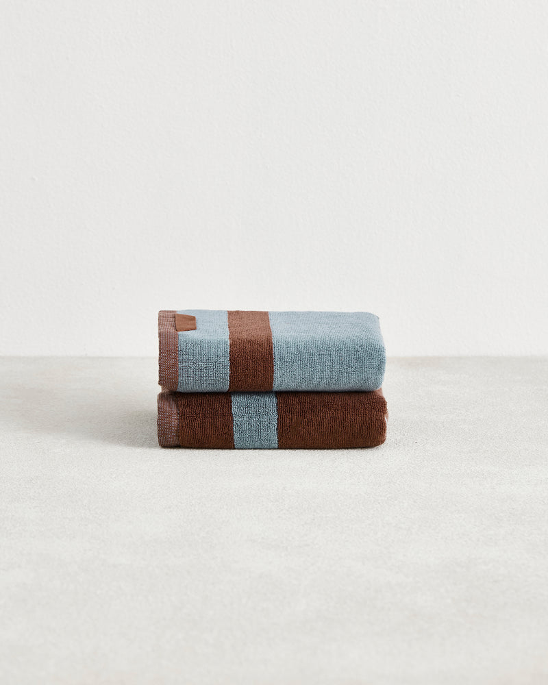 Two Bed Threads Cacao & Lagoon Two-Toned 100% Cotton Terry Hand Towels, featuring plush 650GSM fabric in light blue and brown, are neatly folded and stacked on a light surface against a plain white background. Set of two.