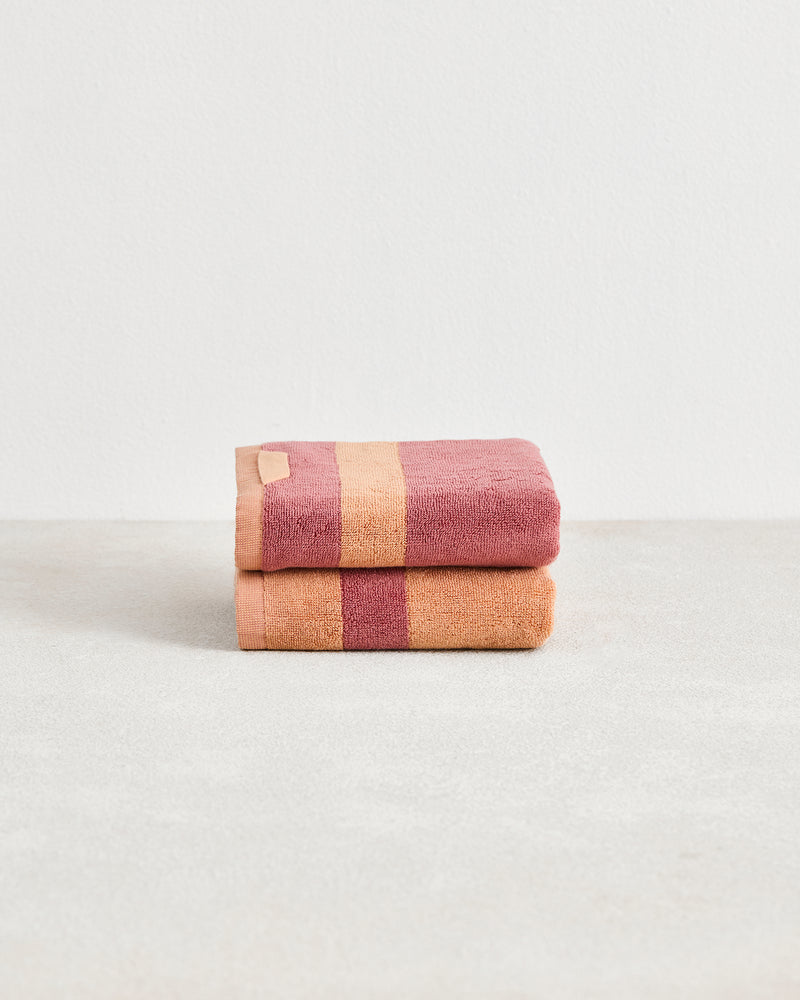 A neatly folded set of two Bed Threads Terracotta & Pink Clay Two-Toned 100% Cotton Terry Hand Towels is stacked on a smooth surface, displaying wide stripes in soft hues against a plain white background.