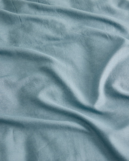 The Lagoon 100% French Flax Linen Flat Sheet by Bed Threads features light blue linen with soft folds and natural creases, offering a smooth, textured surface that helps regulate temperature.