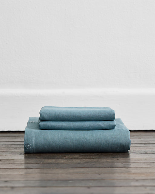 The Lagoon 100% French Flax Linen Duvet Cover Set by Bed Threads, neatly folded and stacked on a wooden floor against a white wall.