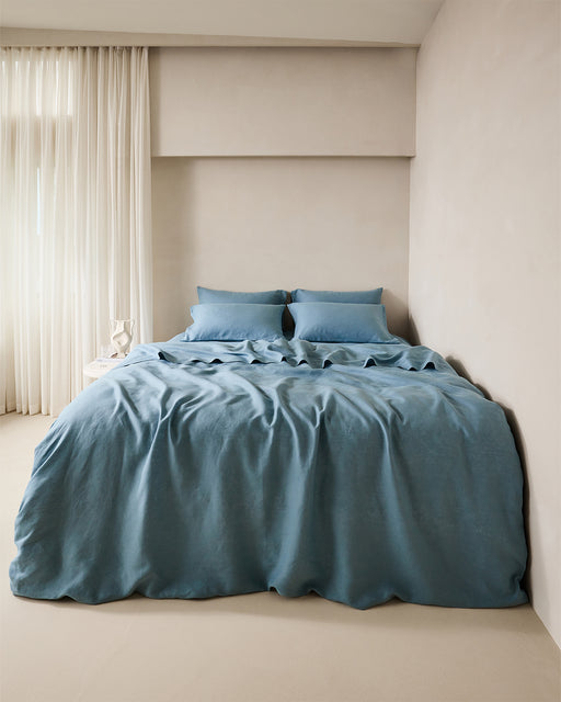 A neatly made bed features the Lagoon 100% French Flax Linen Flat Sheet by Bed Threads and two pillows, set against a light wall. Sheer white curtains cover a window to the left, with a small white side table holding decor nearby.