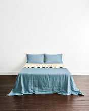 A bed dressed in a dusty blue quilt cover in Lagoon & Creme, with two matching pillowcases in Lagoon, set against a plain white wall and dark wood floor. - view